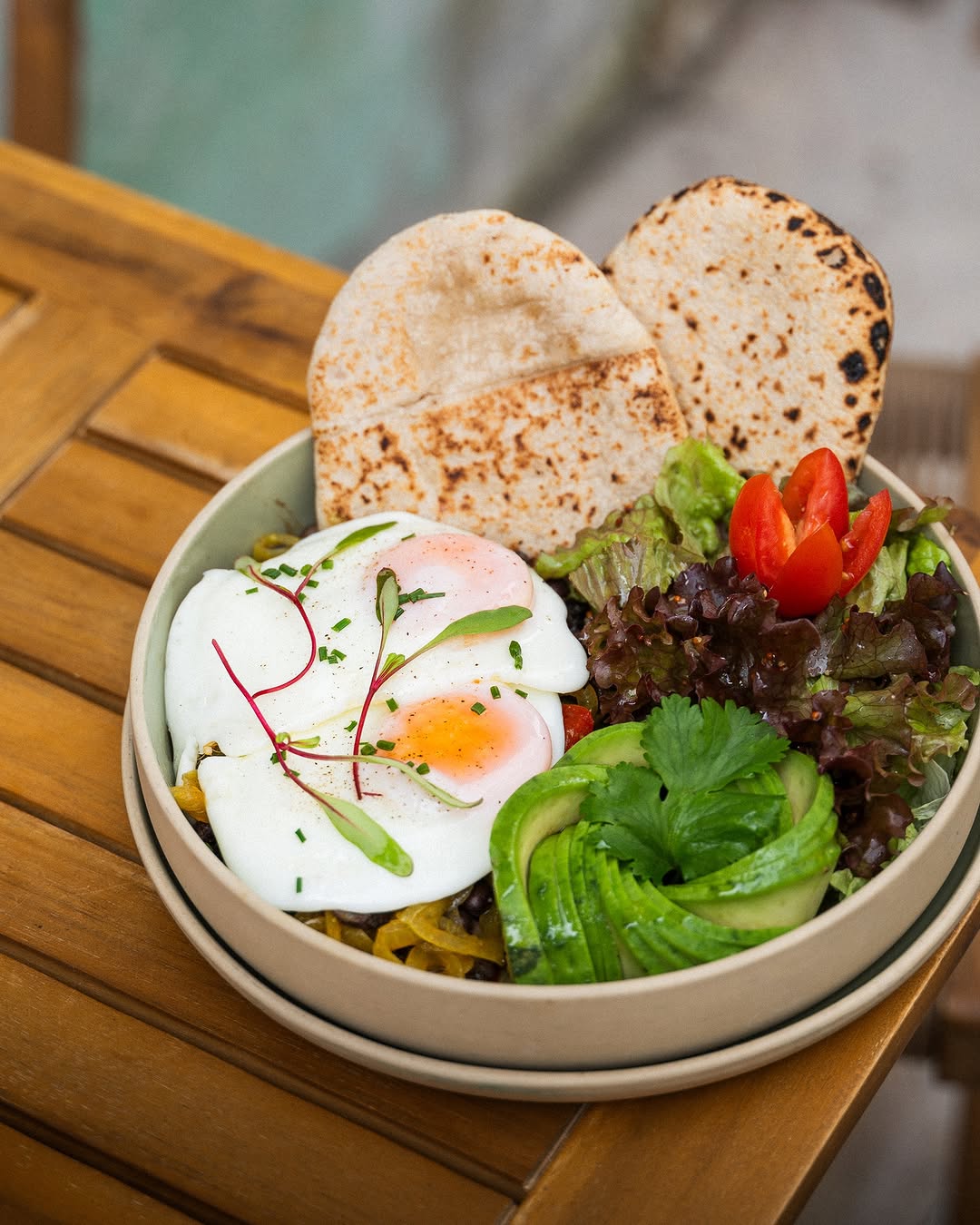 A shallow, round, off-white ceramic bowl containing two poached eggs garnished with microgreens, a mixed green salad with avocado, cherry tomatoes, and leafy greens, and two pieces of toasted flatbread. The food is arranged neatly in the bowl. The setting is a light brown wooden table with visible grain and planks.