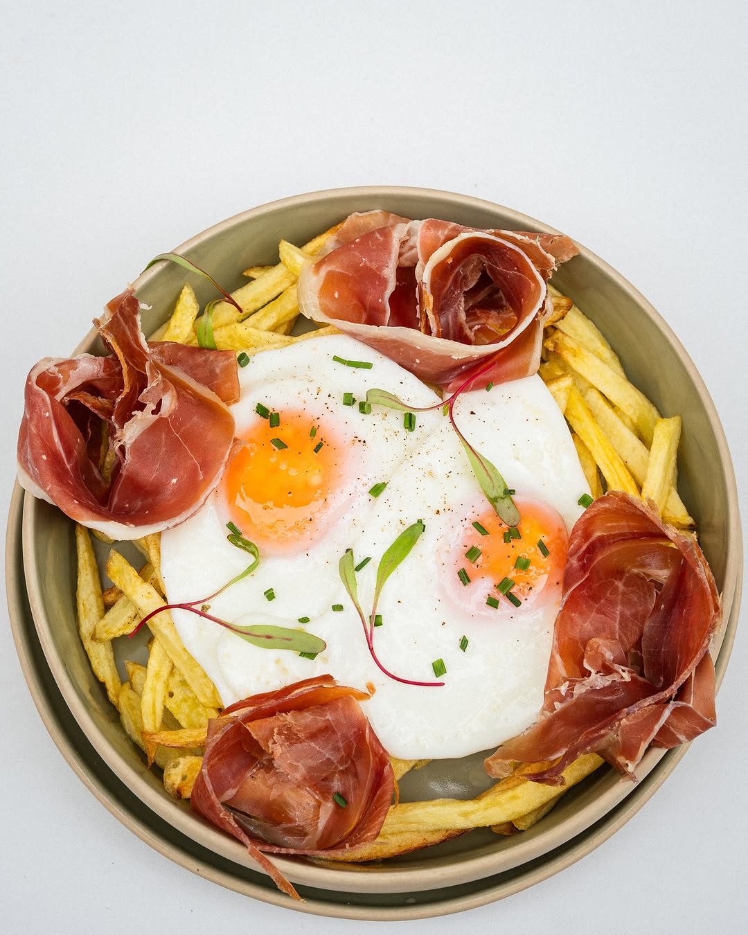 A plate of french fries topped with two fried eggs cooked sunny-side up. Prosciutto is arranged around the eggs, and fresh chives garnish the dish. The plate is light brown and sits on a plain white surface. The food items are the primary focus of the image.