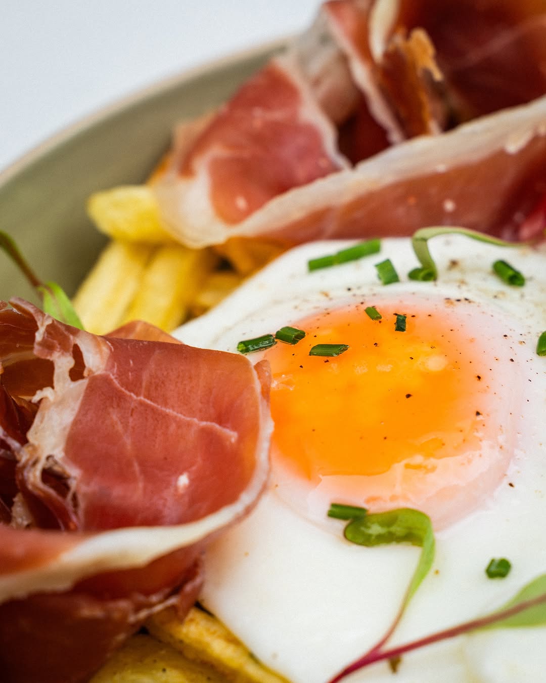 Close-up of a fried egg nestled between slices of prosciutto and golden fries, garnished with chives. The food items are the primary focus of the image.